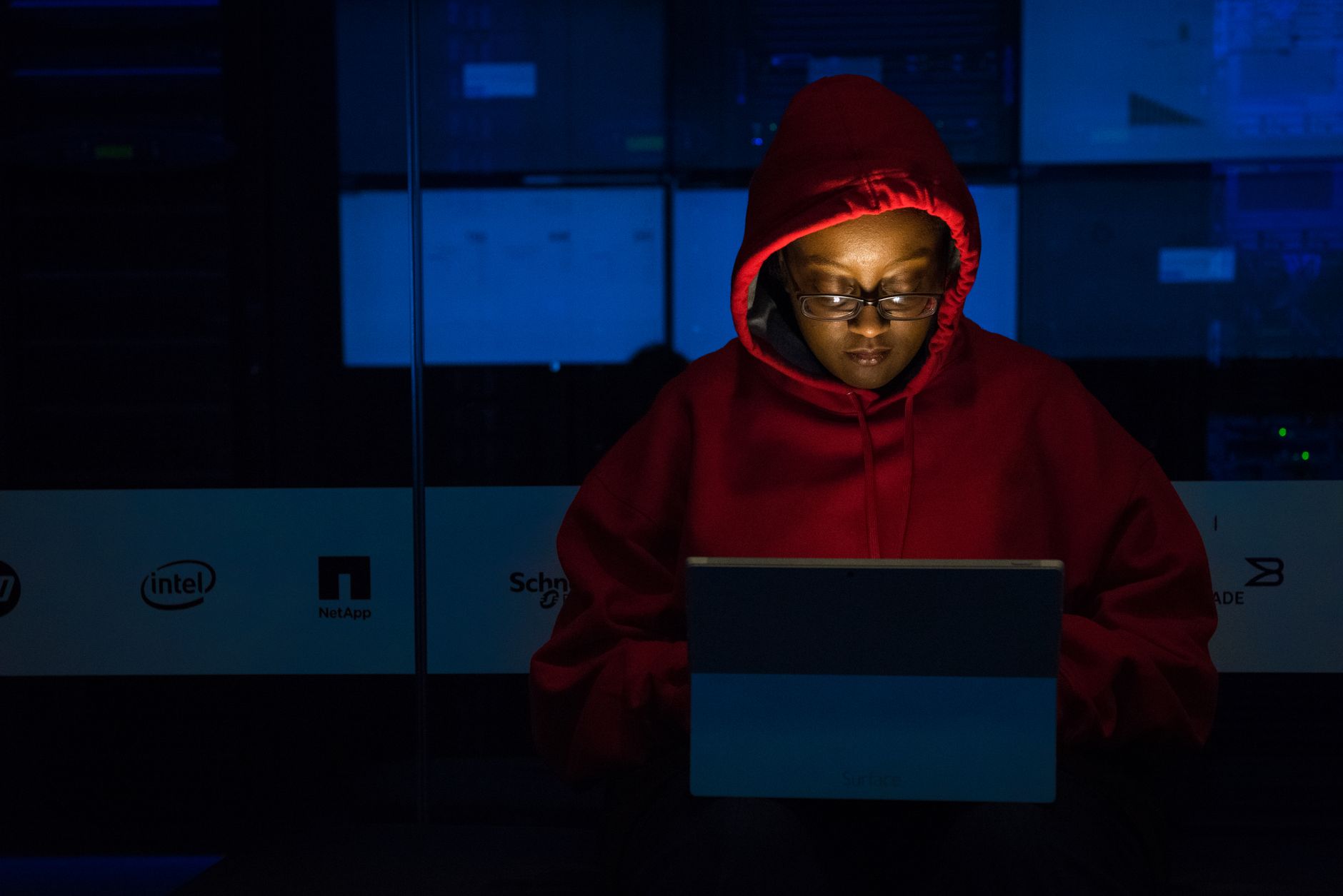 A woman in red hoodie works intensely on her laptop indoors, illuminated by screen light.