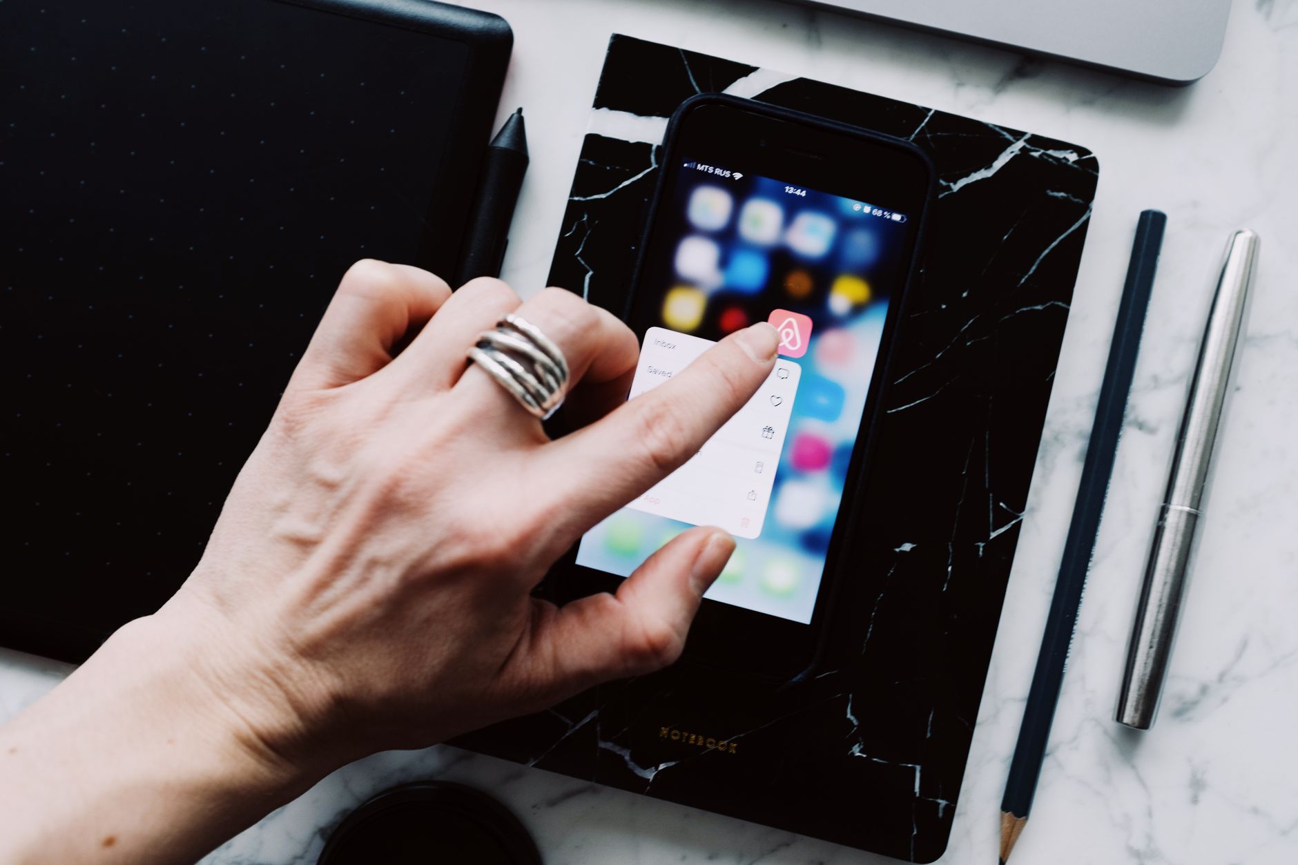 From above of crop faceless person touching cellphone screen with finger to demonstrate app while having gadget near pen and pencil located on marble table