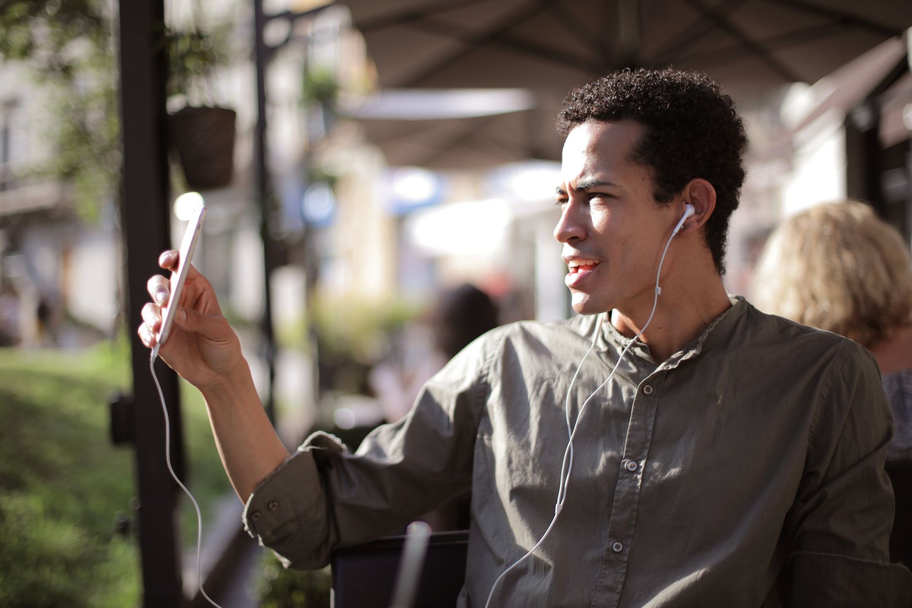 Homme en chemise kaki assis en terrasse, écouteurs aux oreilles, tenant un smartphone devant lui pour regarder l’écran.