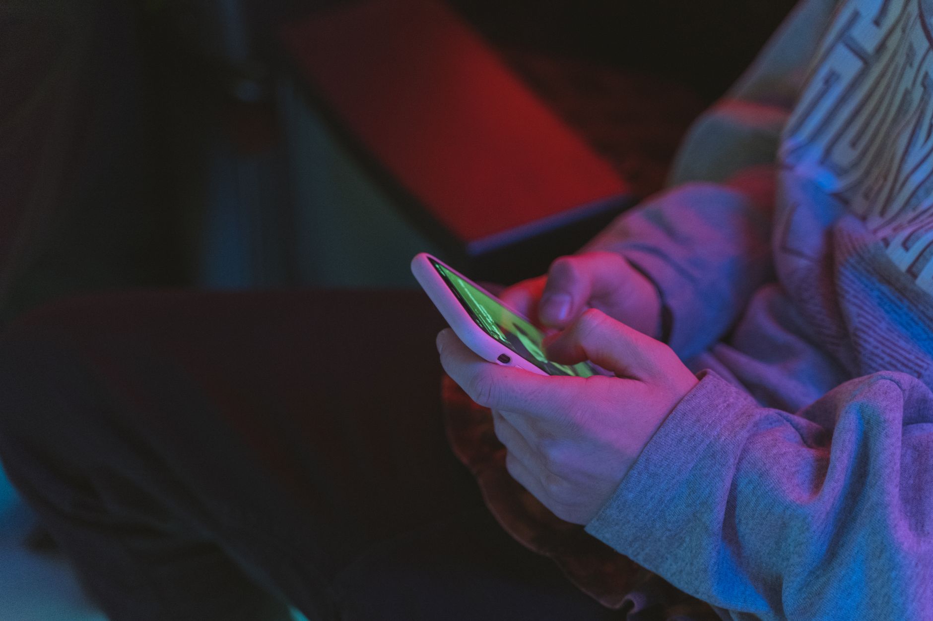 Person in a gray sweatshirt holding a smartphone with a glowing green screen in dim pink and blue light
