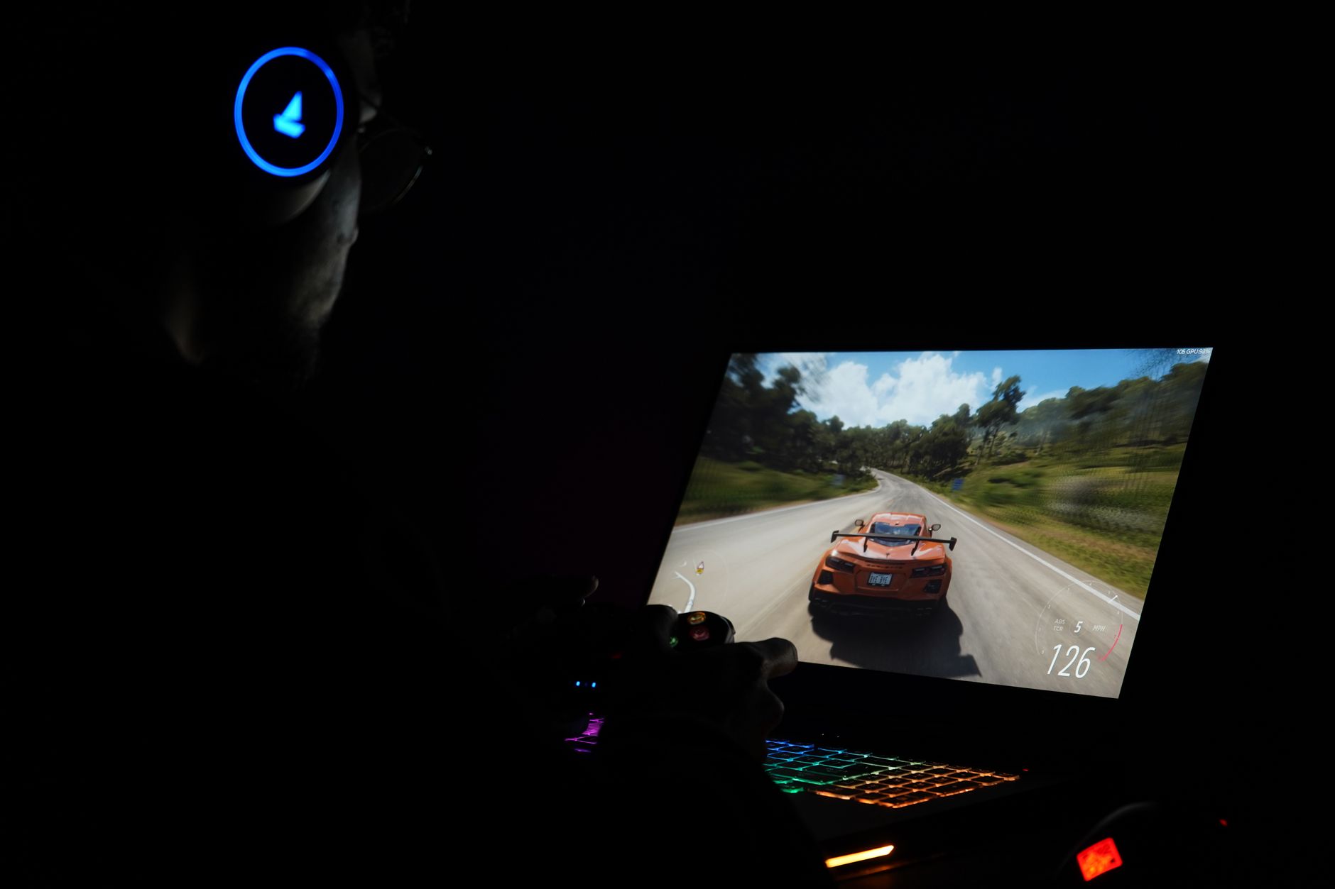Person in headphones playing a racing game on a laptop with a backlit keyboard in a dark room