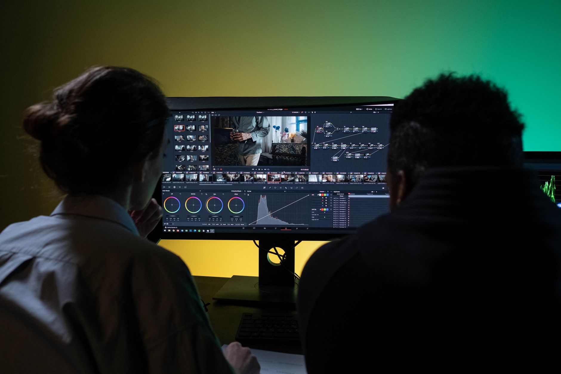 Two people seen from behind look at a wide monitor showing a video editing and color grading interface with a timeline and color wheels.